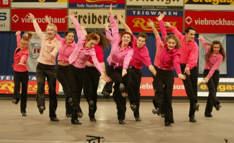 Une troupe de danseurs de claquettes en chemise rose et pantalon noir, bras levés, en pleine chorégraphie sur scène
